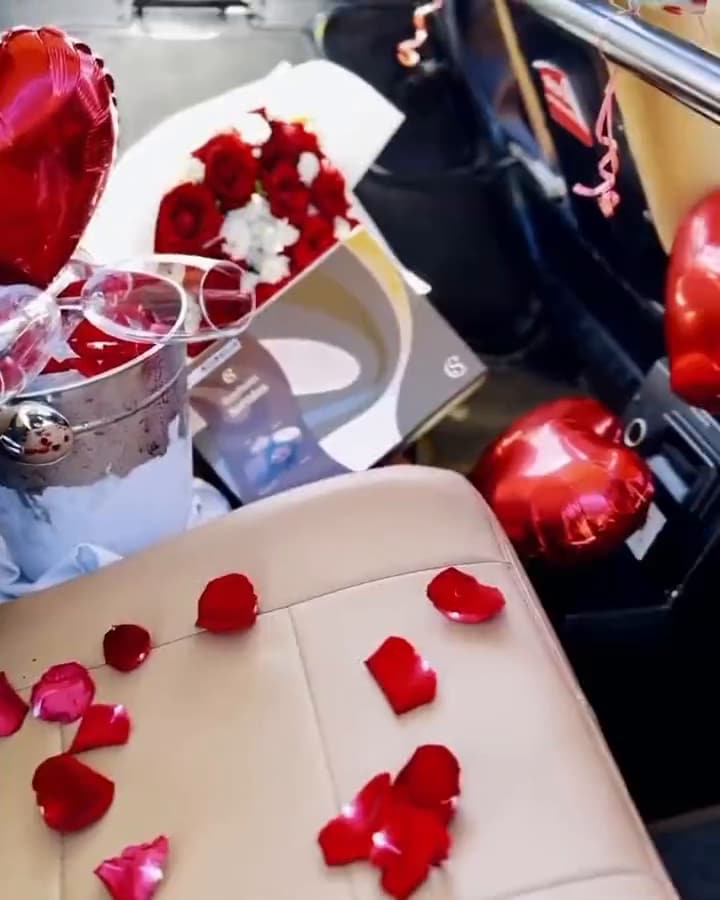 Interior do helicóptero decorado com pétalas de rosa e balões de coração, casal estourando espumante e buquê de rosas vermelhas antes do voo romântico